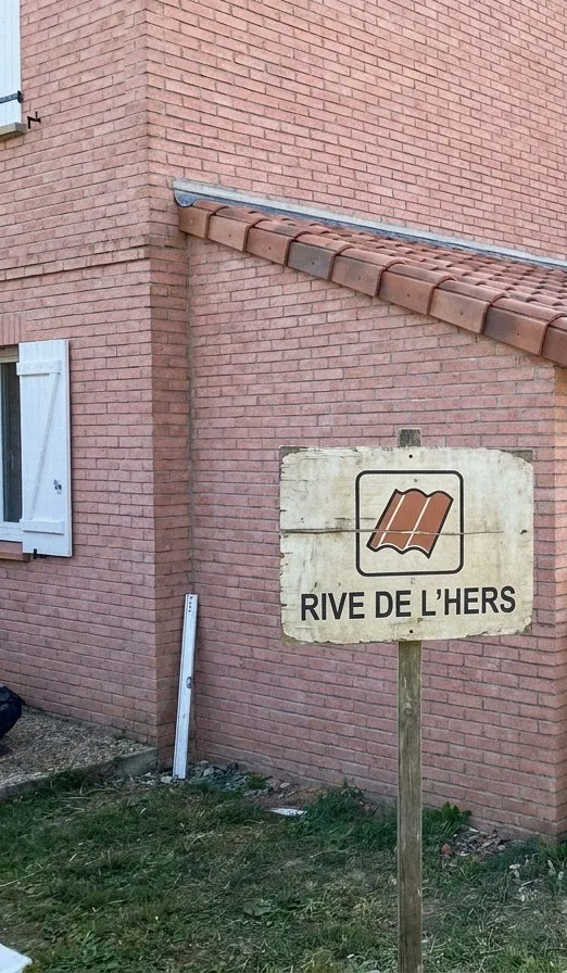 Panneau Rive de l’Hers planté devant une maison en brique rose dont l’appentis vient d’être recouvert en tuiles canal, près de Saint-Jean